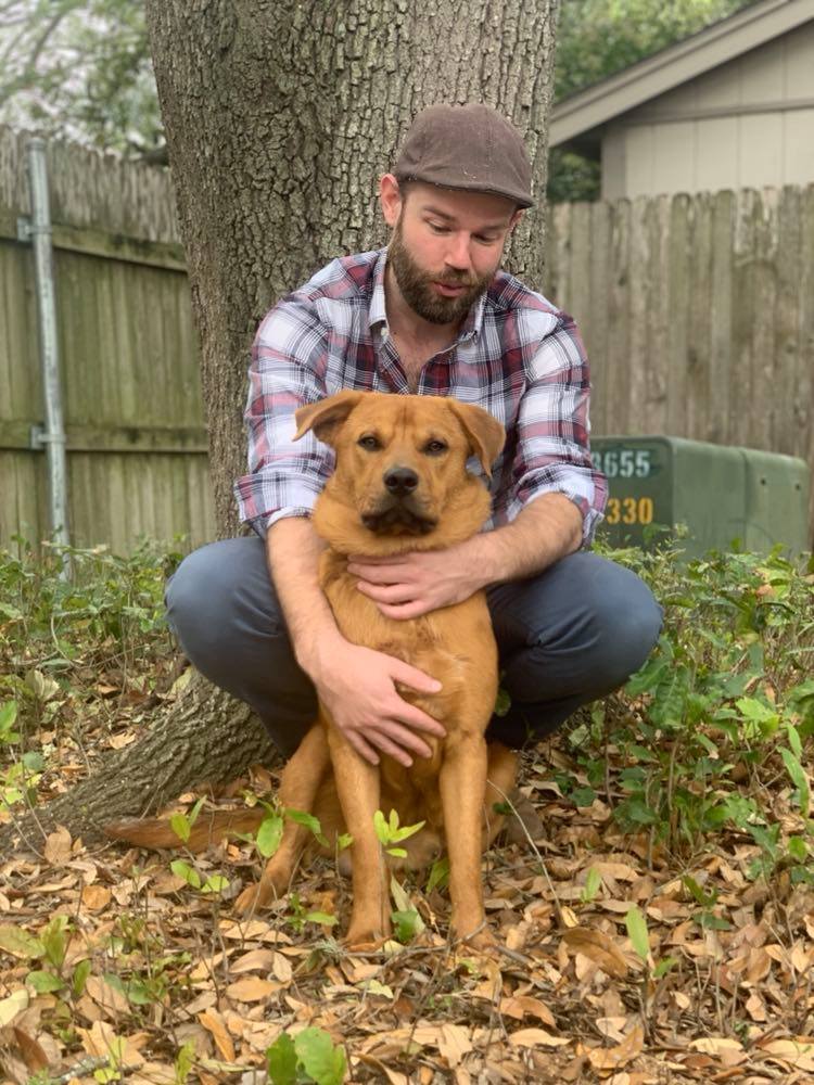 Kye Roper in a flannel shirt and cap hugging a very serious-looking but handsome dog